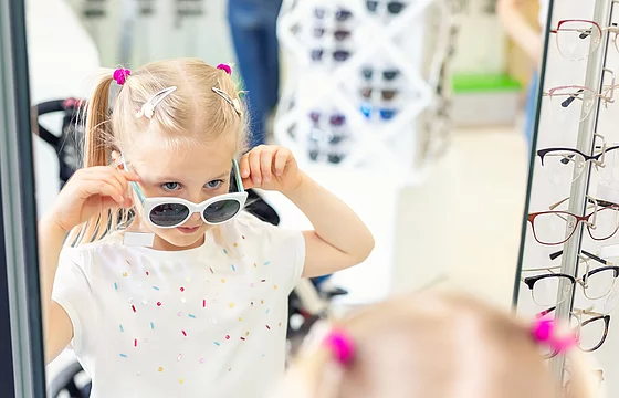 Kind steht mit weiß-blauer Sonnenbrille vor einem Spiegel