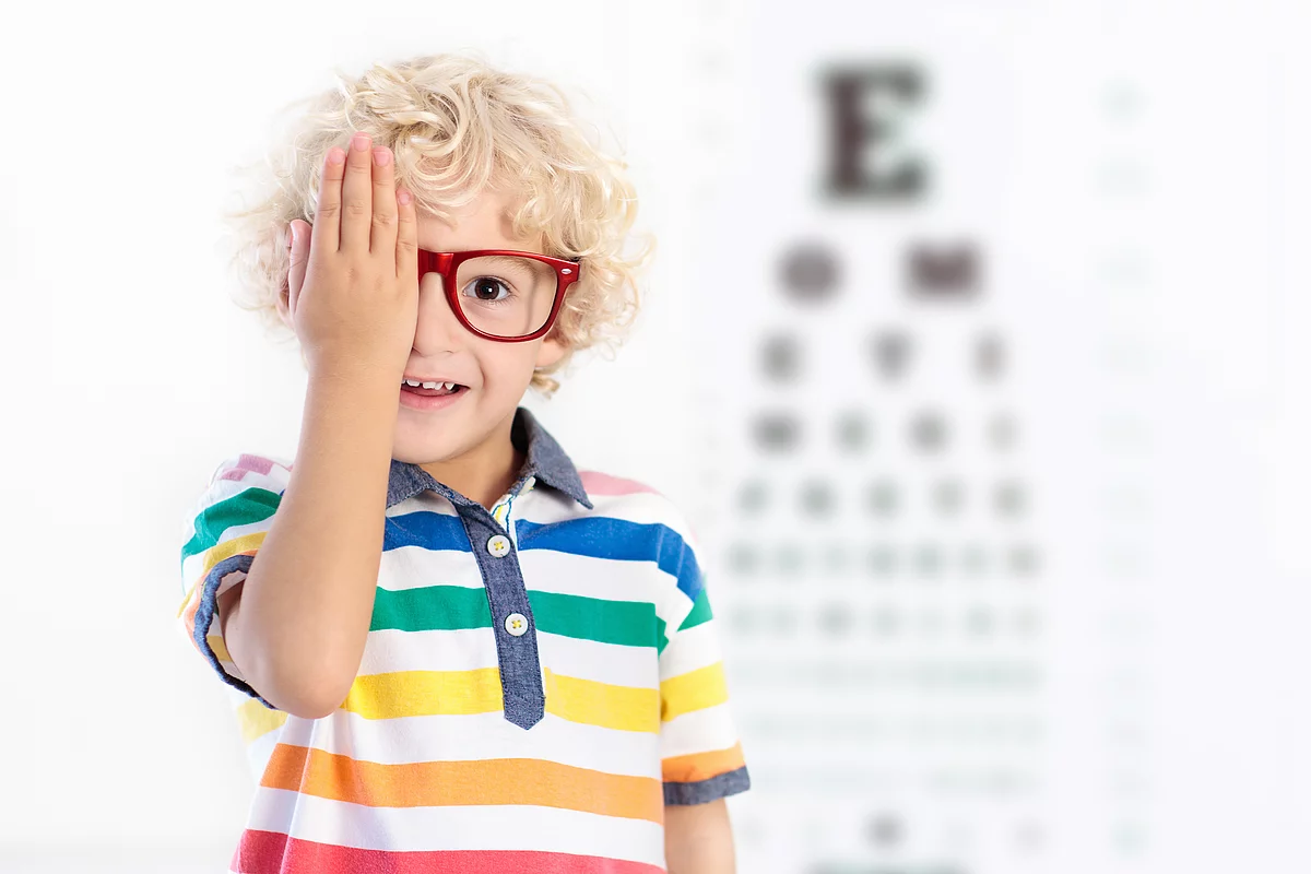 Junge mit roter Brille steht mit dem Rücken vor einer Snellen Tafel und hält sich ein Auge zu